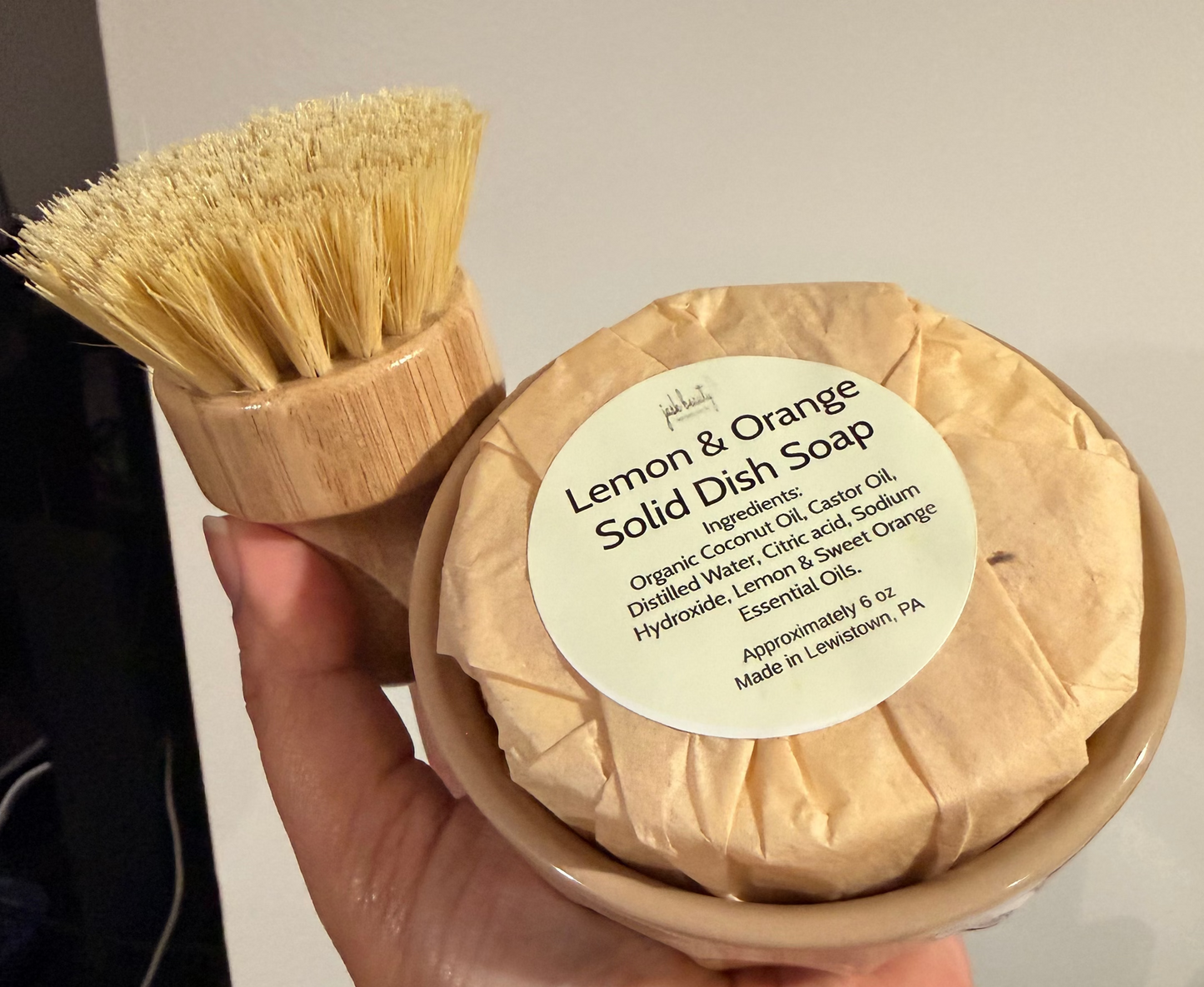 Hand holding a lemon and orange solid dish soap with a brush against a neutral background